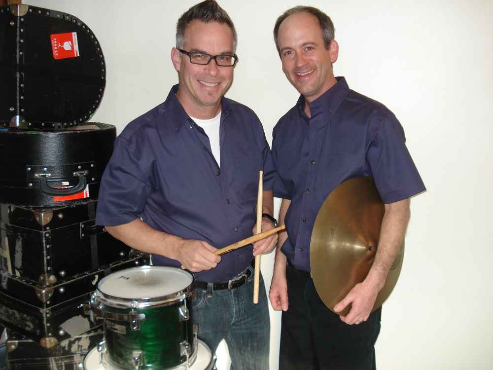 The Rhythm Team: Drummers Charlie Cooley (left) and Jay Boehmer, standing beside a stack of drum cases. Charlie is holding a pair of drumsticks and Jay is holding a cymbal